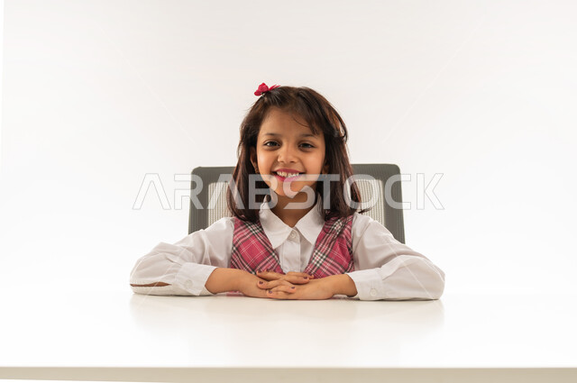 مفهوم العودة الى المدارس, النظر الى الكاميرا بايماءات السعادة والفرح, تلميذه عربيه خليجية عمانية ترتدي الزي المدرسي تستعد لبدء الدرس, بورتريه مقرب لطالبة سعودية تجلس على الطاولة بتعابير السرور, خلفية بيضاء