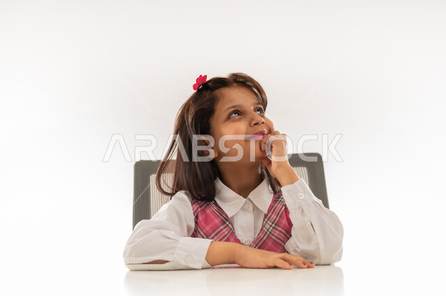 مفهوم العودة الى المدارس, التفكير بالمستقبل بايماءات السعادة والفرح, تلميذه عربيه خليجية عمانية ترتدي الزي المدرسي تضع يدها على ذقنها بتعابير السرور, بورتريه مقرب لطالبة سعودية تجلس على الطاولة تنظر الى الاعلى الى شيء ما, خلفية بيضاء