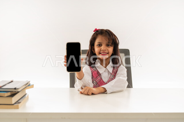 استخدام التقنيات الحديثة في الحياة العلمية، اكاديميات ومدارس متطورة، بورتريه مقرب لتلميذة عربية خليجية عمانية ترتدي الزي المدرسي تقوم بعرض شاشة سوداء فارغه عبر الجوال, طالبة سعودية تجلس على طاولة الدراسة وبجانبها مجموعه من الكتب تنظر الى الكاميرا، خلفية بيضاء