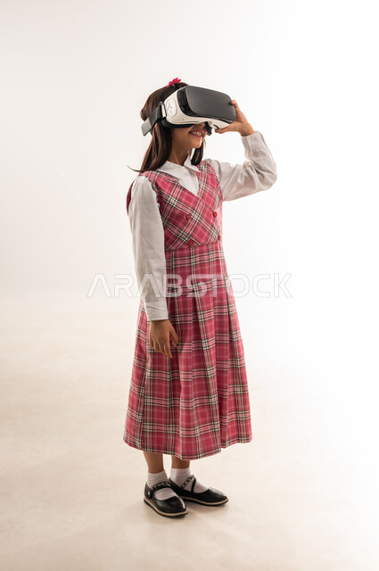 Development of teaching methods using modern advanced technologies, portrait of a Saudi Arabian Gulf girl wearing a school uniform and wearing 3D virtual reality glasses, an Omani schoolgirl looking at something with gestures of integration and concentration, full body length, white background