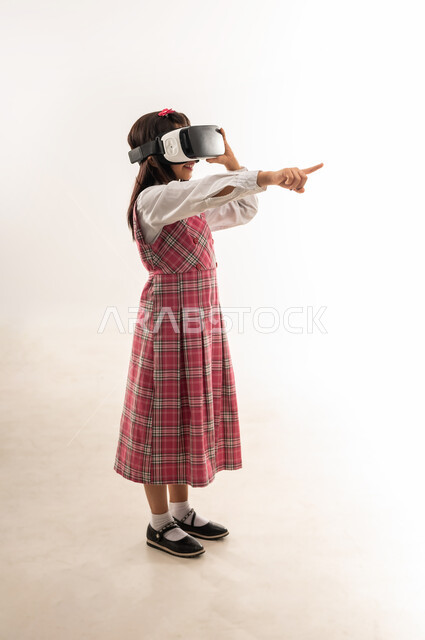 Gestures of integration and concentration, development of teaching methods using modern advanced technologies, portrait of a Saudi Arabian Gulf girl wearing a school uniform and wearing 3D virtual reality glasses, an Omani schoolgirl raising her index finger pointing at something, full body length, white background