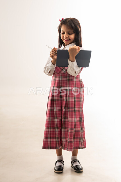 طرق ومناهج تعليمية متطورة، متابعة الدروس عن طريق منصات التعلم عن بعد، مفهوم التعليم المنزلي، تلميذة عمانية تستخدم قلم الايباد لتدوين الملاحظات، بورتريه لطالبة عربية خليجية سعودية ترتدي الزي المدرسي تحمل بيدها جهاز التابلت، الطول الكامل للجسم, خلفية بيضاء