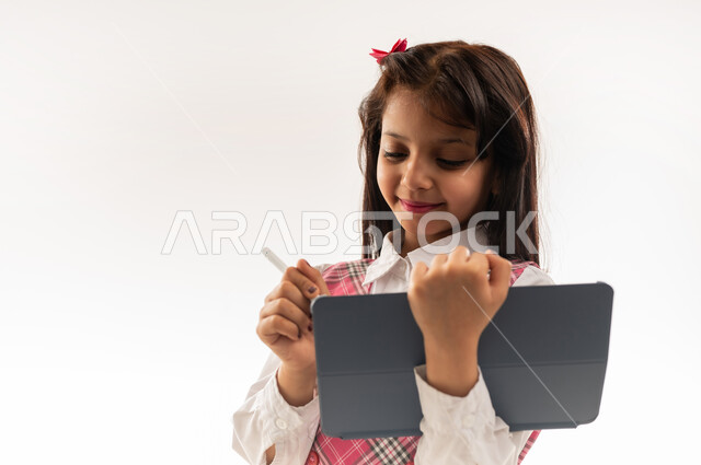 متابعة الدروس عن طريق منصات التعلم عن بعد، مفهوم التعليم المنزلي، تلميذة عمانية تستخدم قلم الايباد لتدوين الملاحظات، بورتريه مقرب لطالبة عربية خليجية سعودية ترتدي الزي المدرسي تحمل بيدها جهاز التابلت التقني، طرق ومناهج تعليمية حديثة ومتطورة، خلفية بيضاء