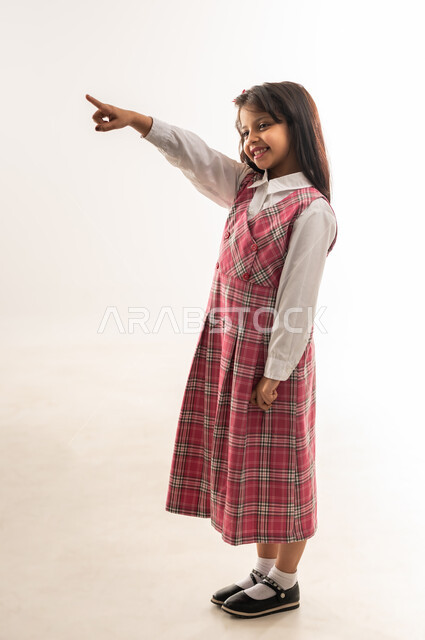 رفع اصبع السبابة نحو الأعلى والاشارة الى هدف ما, بداية العام الدراسي الجديد، تلميذة عمانية تنظر الى شيء ما بتعابير السرور, بورتريه لفتاة سعودية مبتسمة ترتدي الزي المدرسي تقوم بتحديد شيء معين, الجسم بالطول الكامل, خلفية بيضاء