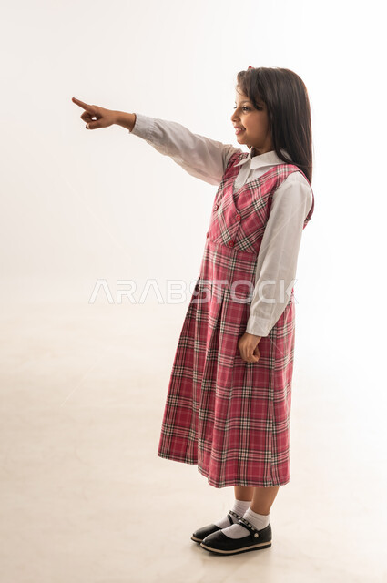 بداية العام الدراسي الجديد، تلميذة عمانية ترفع اصبع السبابة نحو الأعلى تشير لشيء ما بتعابير السرور, بورتريه لفتاة سعودية مبتسمة ترتدي الزي المدرسي تقوم بتحديد شيء معين, الجسم بالطول الكامل, خلفية بيضاء