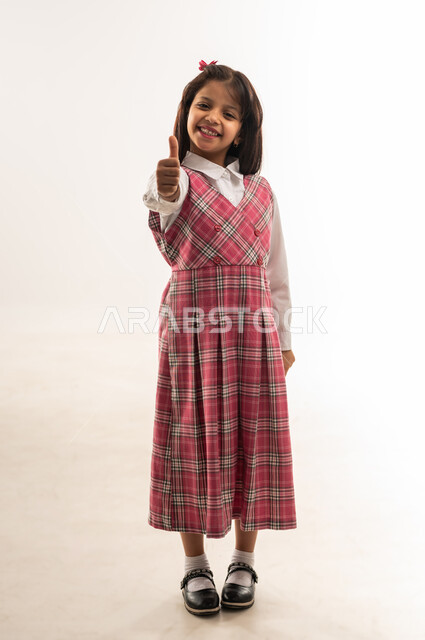 بداية العام الدراسي الجديد، إيماءات الإعجاب بجودة التعليم في دول الخليج, تلميذة عمانية ترفع اصبع الإبهام نحو الأعلى بعلامة النجاح والتفوق, بورتريه لفتاة سعودية مبتسمة ترتدي الزي المدرسي تنظر الى الكاميرا, الطول الكامل للجسم, خلفية بيضاء