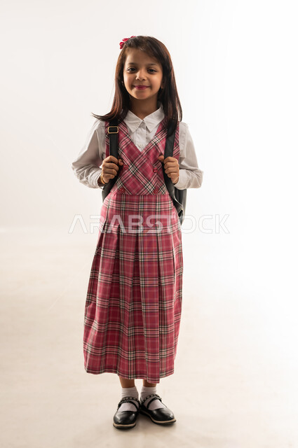 التجهيز و التهيئة لبداية العام الدراسي, تلميذة عمانية تنظر الى الكاميرا بإيماءات السعادة، مفهوم موسم العوده للمدارس, بورتريه لطالبة عربية خليجية سعودية ترتدي اليونيفورم المدرسي تحمل الحقيبة المدرسية, الرغبة في طلب العلم، الجسم بالطول الكامل, خلفية بيضاء