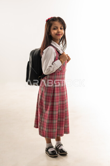 موسم العوده للمدارس, التجهيز لبداية العام الدراسي, بورتريه من الجانب لطالبة عربية خليجية عمانية ترتدي اليونيفورم المدرسي تحمل الحقيبة المدرسية, الرغبة في طلب العلم، تلميذه سعودية تنظر الى الكاميرا بتعابير السرور, الطول الكامل للجسم، خلفية بيضاء
