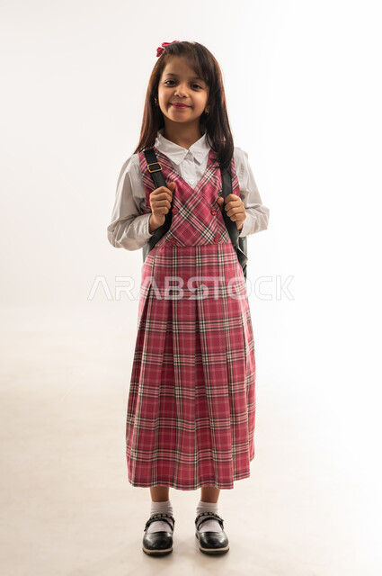 الرغبة في طلب العلم، التهيئة والتجهيز لبداية العام الدراسي, تلميذة عمانية تنظر الى الكاميرا بإيماءات السعادة، مفهوم موسم العوده للمدارس, بورتريه لطالبة عربية خليجية سعودية ترتدي اليونيفورم المدرسي تحمل الحقيبة المدرسية, الجسم بالطول الكامل, خلفية بيضاء