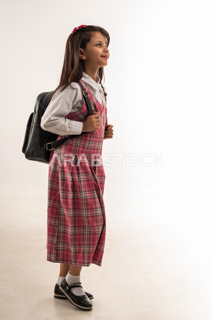 المثابرة لتحقيق التفوق والنجاح، ايماءات السير والتوجه الى المدرسة، بورتريه من الجانب لطالبة عربية خليجية سعودية ترتدي الزي المدرسي وتحمل حقيبة الظهر المدرسية، تلميذة عمانية تستعد لبدء العام الدراسي الجديد بتعابير السرور، الطول الكامل للجسم، خلفية بيضاء