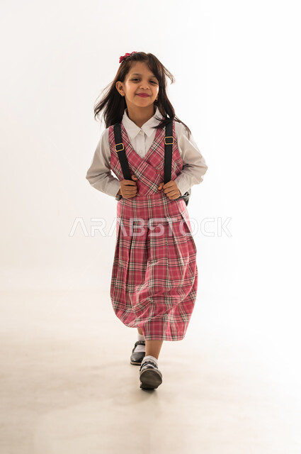 ايماءات السير والتوجه الى المدرسة، بورتريه لطالبة عربية خليجية سعودية ترتدي الزي المدرسي وتحمل حقيبة الظهر المدرسية، تلميذة عمانية تستعد لبدء العام الدراسي الجديد بتعابير السرور، المثابرة لتحقيق التفوق والنجاح، الطول الكامل للجسم، خلفية بيضاء