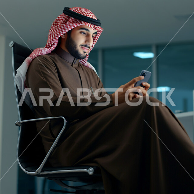 تصفح وسائل التواصل الاجتماعي, استخدام الأجهزة التقنية الحديثة في الحياة اليومية, الدردشة مع العائلة والأصدقاء, شاب عربي خليجي سعودي يرتدي الثوب التقليدي والشماغ يجلس على الكرسي ويمسك بيده الهاتف المحمول