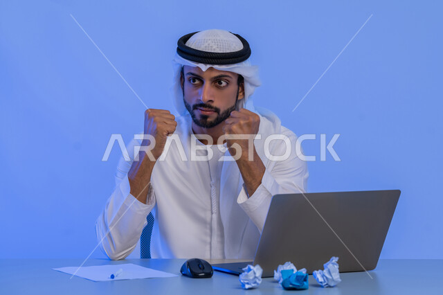 الفشل في العمل، البحث عن افكار وخطط، الاخفاق في الكتابة والتدوين ، مهن ووظائف ادارية مكتبية، بورتريه لرجل عربي خليجي اماراتي يرتدي الكندورة والغترة البيضاء يستخدم الحاسوب المحمول وبجانبه مجموعة من الاوراق المجعدة، استخدام الاجهزة التقنية الحديثة، خلفية زرقاء