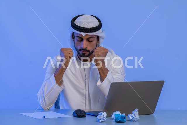 الفشل في العمل، البحث عن افكار وخطط، الاخفاق في الكتابة والتدوين ، مهن ووظائف ادارية مكتبية، بورتريه لرجل عربي خليجي اماراتي يرتدي الكندورة والغترة البيضاء يستخدم الحاسوب المحمول وبجانبه مجموعة من الاوراق المجعدة، استخدام الاجهزة التقنية الحديثة، خلفية زرقاء