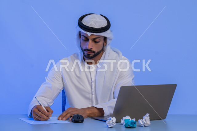الفشل في العمل، البحث عن افكار وخطط، الاخفاق في الكتابة والتدوين ، مهن ووظائف ادارية مكتبية، بورتريه لرجل عربي خليجي اماراتي يرتدي الكندورة والغترة البيضاء يستخدم الحاسوب المحمول وبجانبه مجموعة من الاوراق المجعدة، استخدام الاجهزة التقنية الحديثة، خلفية زرقاء
