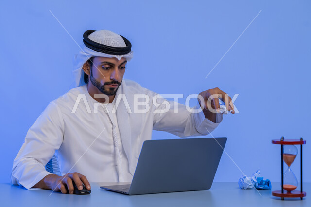 استخدام الساعة الرملية لتحديد موعد العمل، مهن ووظائف ادارية مكتبية في الامارات، مفهوم إدارة شؤون الأعمال وتنظيمها عبر الاجهزة التقنية، بورتريه مقرب لرجل عربي خليجي اماراتي يرتدي الكندورة والغترة البيضاء يقوم باستخدام جهاز الحاسوب المحمول، خلفية زرقاء