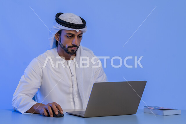 مهن ووظائف ادارية مكتبية في الامارات، مفهوم إدارة شؤون الأعمال وتنظيمها عبر الاجهزة التقنية، بورتريه مقرب لرجل أعمال عربي خليجي اماراتي يرتدي الكندورة والغترة البيضاء يقوم باستخدام جهاز الحاسوب المحمول، استخدام التقنيات الحديثة والمتطورة، خلفية زرقاء