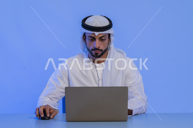 مهن ووظائف ادارية مكتبية في الامارات، مفهوم إدارة شؤون الأعمال وتنظيمها عبر الاجهزة التقنية، بورتريه مقرب لرجل أعمال عربي خليجي اماراتي يرتدي الكندورة والغترة البيضاء يقوم باستخدام جهاز الحاسوب المحمول، استخدام التقنيات الحديثة والمتطورة، خلفية زرقاء