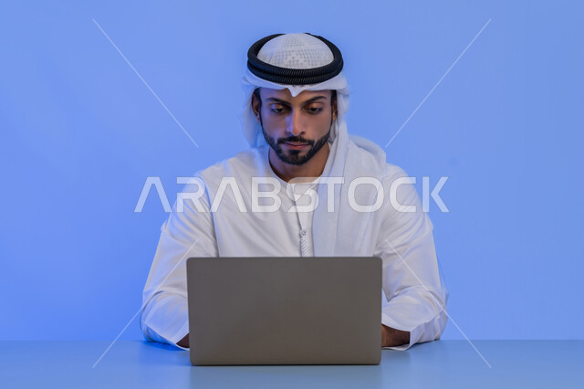 مهن ووظائف ادارية مكتبية في الامارات، مفهوم إدارة شؤون الأعمال وتنظيمها عبر الاجهزة التقنية، بورتريه مقرب لرجل أعمال عربي خليجي اماراتي يرتدي الكندورة والغترة البيضاء يقوم باستخدام جهاز الحاسوب المحمول، استخدام التقنيات الحديثة والمتطورة، خلفية زرقاء