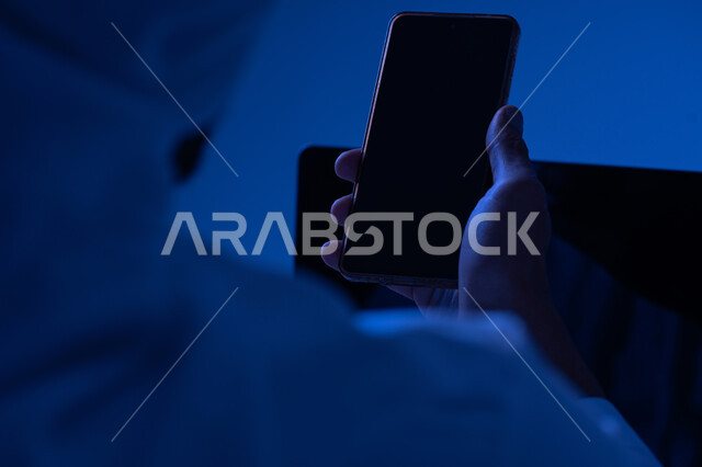 استخدام الاجهزة التقنية الحديثة، صورة مقربة من الخلف لرجل عربي خليجي اماراتي يحمل بيده الجوال وبجانبه جهاز اللابتوب، شاب سعودي يقوم بنقل البيانات والملفات بين الجهازين، عرض شاشة سوداء فارغة عبر الهاتف والحاسوب المحمول، التسويق والترويج للمواقع الالكترونية