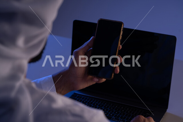 استخدام الاجهزة التقنية الحديثة، صورة مقربة من الخلف لرجل عربي خليجي اماراتي يحمل بيده الجوال وبجانبه جهاز اللابتوب، شاب سعودي يقوم بنقل البيانات والملفات بين الجهازين، عرض شاشة سوداء فارغة عبر الهاتف والحاسوب المحمول، التسويق والترويج للمواقع الالكترونية