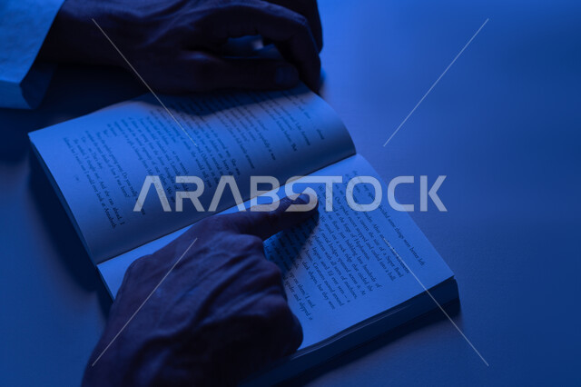 تنمية الذخيرة الثقافية، قضاء أوقات الفراغ بممارسة هواية المطالعة، مفهوم الثقافة والتعلم الذاتي, شاب سعودي يقوم بقراءة القصص والروايات الممتعة, صورة مقربة لأيدي رجل عربي خليجي إماراتي يرتدي الكندورة يقوم بتتبع الكتابة في الكتاب