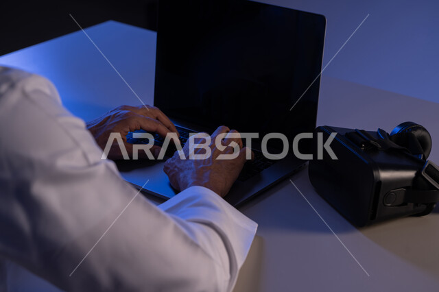 استخدام الأجهزة التكنولوجية والتقنيات الحديثة, محاكاة العالم الخيالي, صورة مقربة من الخلف لرجل عربي خليجي إماراتي يقوم بإنجاز العمل على الحاسوب المحمول وبجانبه نظارة الفي ار, شاب سعودي يمتلك مهارة الكتابة السريعة على الكيبورد