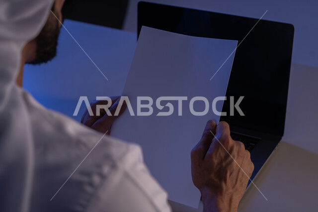 قراءة الملاحظات والمعلومات، متابعة الخطط والمشاريع، صورة مقربة من الخلف لرجل عربي خليجي إماراتي يحمل بيده مجموعة من الاوراق الفارغة ويستخدم الحاسوب المحمول، شاب سعودي يدقق الوثائق الخاصة بالعمل، استحدام الاجهزة التقنية الحديثة والمتطورة