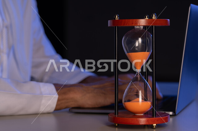 ادارة الجدول الزمني، مفهوم تنظيم الوقت، صورة مقربة لأيدي رجل عربي خليجي إماراتي يرتدي الكندورة يقوم بالكتابة على لوحة مفاتيح الحاسب المحمول، استخدام الساعة الرملية لتحديد موعد العمل، شاب سعودي يقوم بانجاز المهام بدقة وكفاءة والوقت المناسب