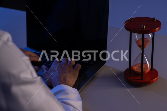 مفهوم تنظيم الوقت، صورة مقربة لأيدي رجل عربي خليجي إماراتي يرتدي الكندورة يقوم بالكتابة على لوحة مفاتيح الحاسب المحمول، استخدام الساعة الرملية لتحديد موعد العمل، شاب سعودي يقوم بانجاز المهام بدقة وكفاءة والوقت المناسب، ادارة الجدول الزمني