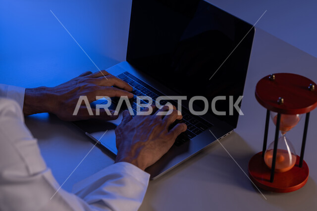 استخدام الساعة الرملية لتحديد موعد العمل، شاب سعودي يقوم بانجاز المهام بدقة وكفاءة والوقت المناسب، ادارة الجدول الزمني، مفهوم تنظيم الوقت، صورة مقربة لأيدي رجل عربي خليجي إماراتي يرتدي الكندورة يقوم بالكتابة على لوحة مفاتيح الحاسب المحمول