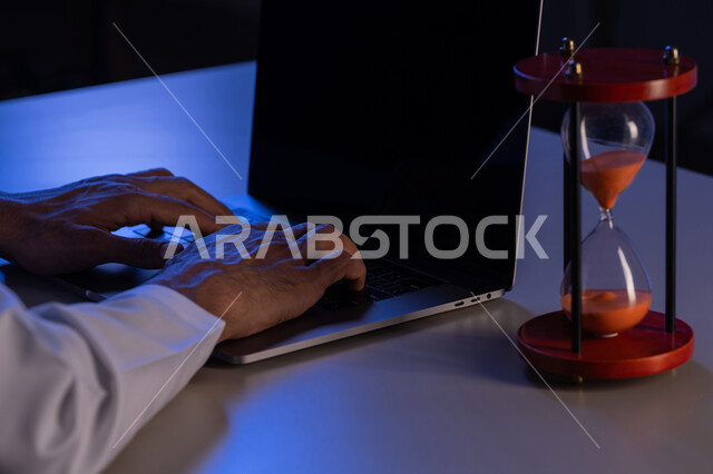 مفهوم تنظيم الوقت، صورة مقربة لأيدي رجل عربي خليجي إماراتي يرتدي الكندورة يقوم بالكتابة على لوحة مفاتيح الحاسب المحمول، استخدام الساعة الرملية لتحديد موعد العمل، شاب سعودي يقوم بانجاز المهام بدقة وكفاءة والوقت المناسب، ادارة الجدول الزمني