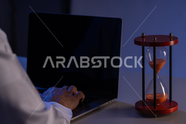 استخدام الساعة الرملية لتحديد موعد العمل، شاب سعودي يقوم بانجاز المهام بدقة وكفاءة والوقت المناسب، ادارة الجدول الزمني، مفهوم تنظيم الوقت، صورة مقربة لأيدي رجل عربي خليجي إماراتي يرتدي الكندورة يقوم بالكتابة على لوحة مفاتيح الحاسب المحمول