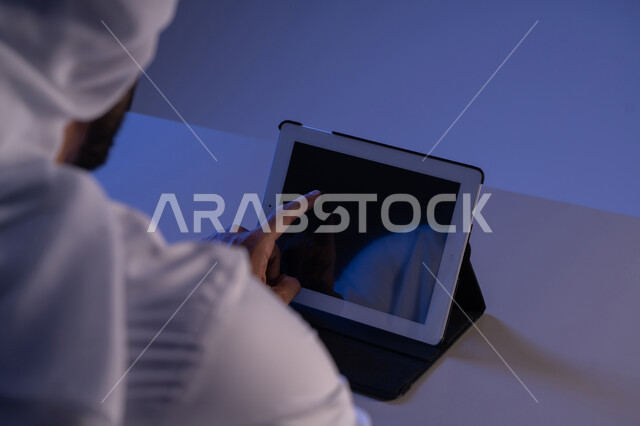 Using modern and advanced technical devices, following educational lessons via tablet, displaying a blank black screen, a close-up image from the back of an Arab Gulf Emirati man completing the required tasks remotely, a Saudi young man using an iPad, browsing social media sites