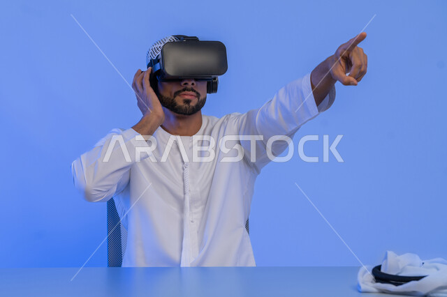 الاشارة إلى شيء ما بأصبع السبابة، الاستمتاع في قضاء الأوقات الترفيهية عبر استخدام التقنيات الحديثة الالكترونية، بورتريه مقرب لشاب عربي خليجي إماراتي يرتدي الكندورة والغترة يضع نظارات الفي آر ثلاثية الابعاد، الاندماج والتفاعل مع العالم الافتراضي، خلفية زرقاء