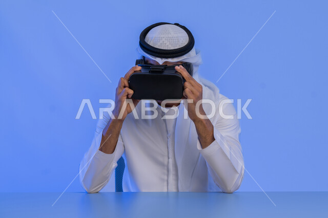 الاندماج والتفاعل مع العالم الافتراضي، دمج التكنولوجيا بالحياة العملية، الاستمتاع في قضاء الأوقات الترفيهية عبر استخدام التقنيات الحديثة الالكترونية، بورتريه مقرب لشاب عربي خليجي إماراتي يرتدي الكندورة والغترة يضع نظارات الفي آر ثلاثية الابعاد، خلفية زرقاء