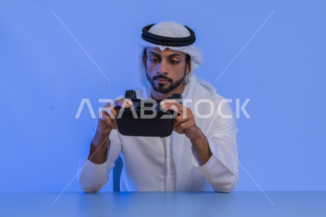 الاستمتاع في قضاء الأوقات الترفيهية عبر استخدام التقنيات الحديثة الالكترونية، بورتريه مقرب لشاب عربي خليجي إماراتي يرتدي الكندورة والغترة يقوم بارتداء نظارات الفي آر ثلاثية الابعاد، الاندماج والتفاعل مع العالم الافتراضي، دمج التكنولوجيا بالحياة العملية، خلفية زرقاء