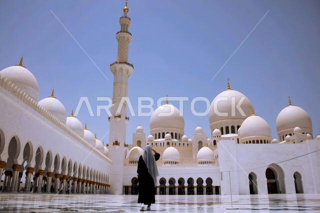 التصاميم الهندسية المميزة للقبب والمآذن والأعمدة البيضاء، صورة من الخلف لإمرأة عربية خليجية اماراتية محجبة ترتدي عباءة سوداء تقف في الساحة الخارجية لمسجد الشيخ زايد الكبير في الامارات العربية المتحدة، الأماكن الإسلامية المقدسة، المعالم المشهورة في مدينة ابو ظبي