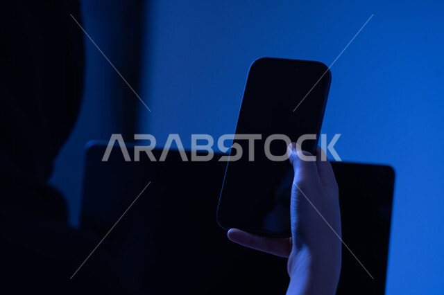 استخدام الاجهزة التقنية الحديثة، صورة مقربة من الخلف لامرأة عربية خليجية اماراتية تحمل بيدها الهاتف المحمول، شابة سعودية تقوم بالعمل على جهاز اللابتوب، عرض شاشة سوداء فارغة، التسويق والترويج للمواقع الالكترونية