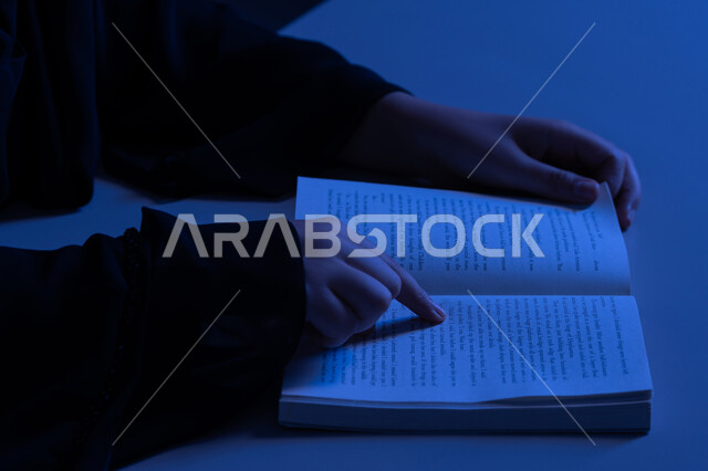 ممارسة هواية المطالعة، الاستمتاع بالأنشطة الترفيهية المفيدة في وقت الفراغ، مفهوم الثقافة والتعلم الذاتي, شابة سعودية تقوم بقراءة القصص والروايات الممتعة, صورة مقربة لأيدي امرأة عربية خليجية اماراتية ترتدي العباءة تقوم بتتبع السطور في الكتاب