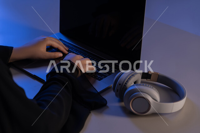 مفهوم الإصغاء والاستماع، شابة سعودية تستخدام التكنولوجيا الحديثة، استعمال تقنية الهيدفون، قضاء أوقات ممتعة بسماع الاغاني والمقاطع الصوتية، عرض شاشة سوداء فارغة عبر الحاسوب المحمول، صورة مقربة لايدي امرأة عربية خيجية اماراتية ترتدي العباءة تقوم بتدوين الملاحظات