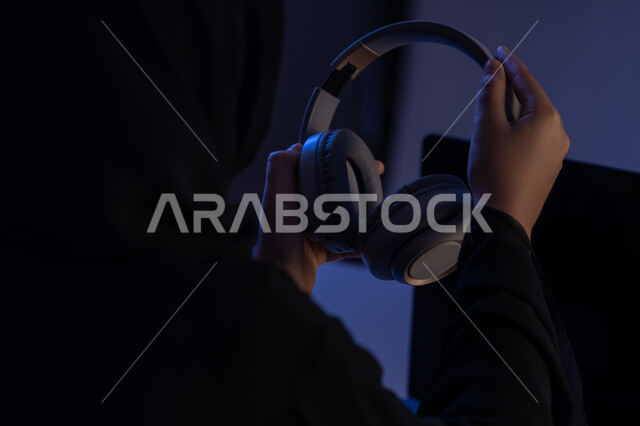 استعمال تقنية الهيدفون، مفهوم الإصغاء والاستماع، شابة سعودية تستخدام التكنولوجيا الحديثة، قضاء أوقات ممتعة بسماع الاغاني والمقاطع الصوتية، صورة مقربة من الجانب لامرأة عربية خيجية اماراتية ترتدي العباءة تقوم بيدها سماعة الرأس
