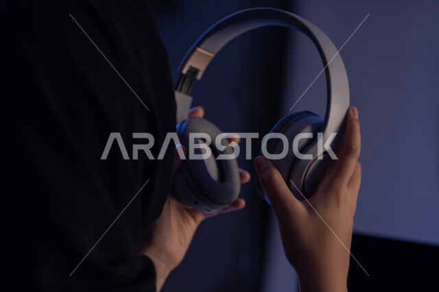 قضاء أوقات ممتعة بسماع الاغاني والمقاطع الصوتية، استعمال تقنية الهيدفون، مفهوم الإصغاء والاستماع، شابة سعودية تستخدام التكنولوجيا الحديثة، صورة مقربة من الجانب لامرأة عربية خيجية اماراتية ترتدي العباءة تقوم بيدها سماعة الرأس