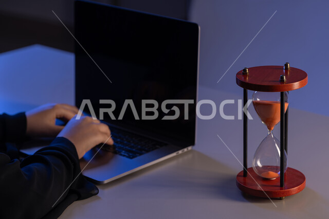 مفهوم تنظيم الوقت، الالتزام بالموعد المحدد في انجاز الاعمال، استخدام الساعة الرملية في ادارة الجدول الزمني، شابة سعودية تقوم بانجاز المهام بدقة وكفاءة، صورة مقربة لأيدي امرأة عربية خليجية اماراتية ترتدي العباءة تقوم بالكتابة على لوحة مفاتيح الحاسب المحمول