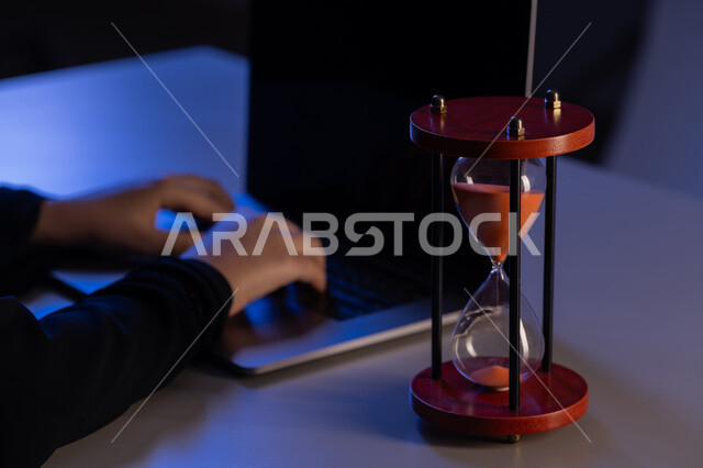 Commitment to deadlines in completing tasks, using hourglass to manage timetable, young Saudi woman completing tasks accurately and efficiently, concept of time management, close-up of hands of Arab Gulf Emirati woman wearing abaya typing on laptop keyboard