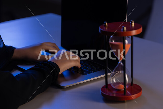 استخدام الساعة الرملية في ادارة الجدول الزمني، شابة سعودية تقوم بانجاز المهام بدقة وكفاءة، الالتزام بالموعد المحدد في انجاز الاعمال، مفهوم تنظيم الوقت، صورة مقربة لأيدي امرأة عربية خليجية اماراتية ترتدي العباءة تقوم بالكتابة على لوحة مفاتيح الحاسب المحمول