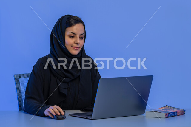 دمج التكنولوجيا الحديثة بالحياة العملية، مفهوم ادخال البيانات، بورتريه مقرب لامرأة عربية خليجية اماراتية تعمل على الحاسوب المحمول، شابة سعودية تجلس امام المكتب، مهن ووظائف إدارية مكتبية، خلفية زرقاء