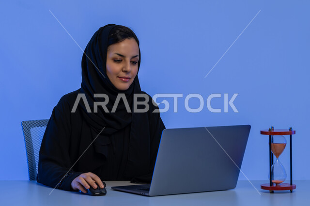 مفهوم تنظيم الوقت، الالتزام بالموعد المحدد في انجاز الاعمال، استخدام الساعة الرملية في ادارة الجدول الزمني، شابة سعودية تقوم بانجاز المهام بدقة وكفاءة، بورتريه مقرب لامرأة عربية خليجية اماراتية ترتدي العباءة تقوم بالكتابة على الحاسب المحمول، خلفية زرقاء