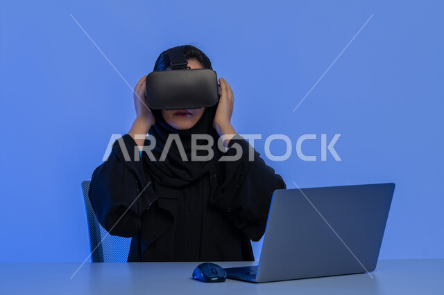 Fantasy world simulation, use of technological devices and modern techniques, close-up portrait of an Arab Gulf Emirati woman doing work on a laptop, a young Saudi woman wearing virtual reality glasses, blue background