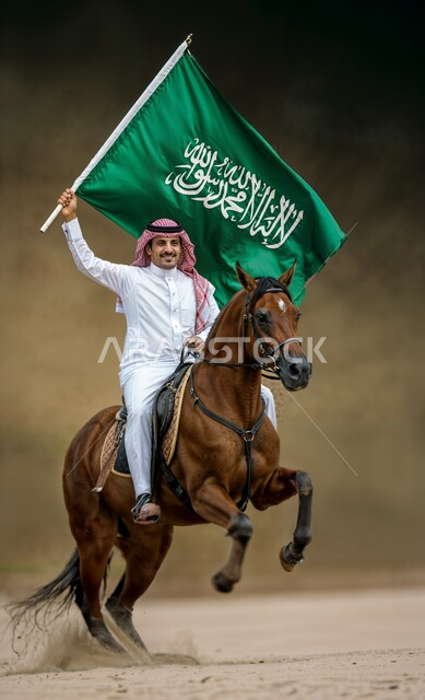 مفهوم الاعتزاز والفخر بالوطن, الاحتفال بالأعياد والمناسبات الوطنية, استذكار اليوم الوطني السعودي 23 سبتمبر, ذكرى يوم العلم 11 مارس, رجل عربي خليجي سعودي يرتدي الثوب التقليدي والشماغ يمتطي حصان بني ويرفع بيده علم المملكة العربية السعودية, خلفية بنية